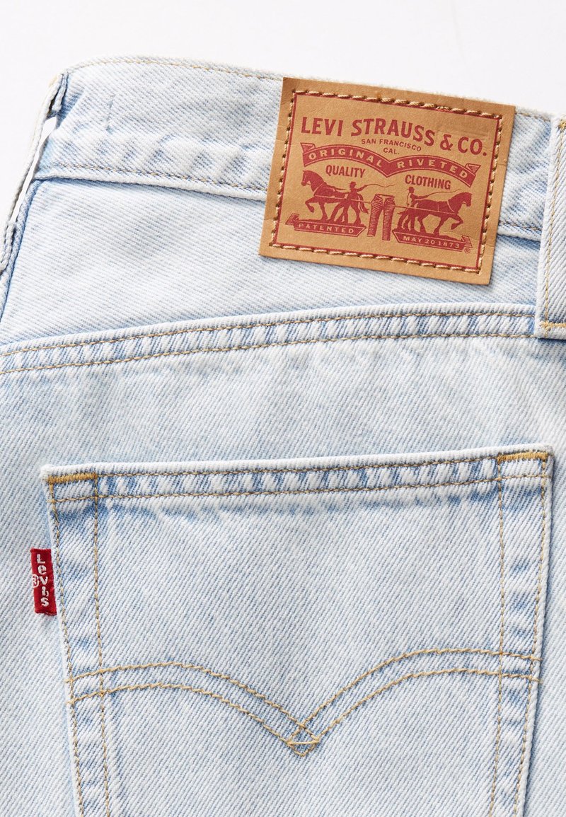 Jeans in denim azzurro chiaro con una toppa in pelle con dettagli rossi, cuciture distintive e un'etichetta rossa sulla tasca posteriore.