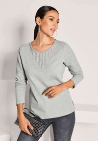 Helles graues Langarmoberteil mit Rundhalsausschnitt, verziert mit Perlenfransen. Kombiniert mit dunklen Jeans und einer schwarzen Clutch mit Perlen.
