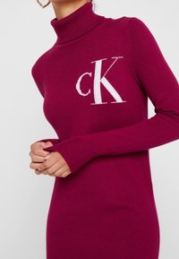 Långärmad klänning med polokrage i klar magentafärgad stickad tyg, med en vit "CK"-logotyp på bröstet och åtsittande ärmslut.