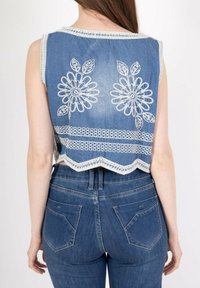 Crop top en denim avec broderie florale blanche au dos, ourlet festonné et bord contrastant. Porté avec un jean bleu taille haute.