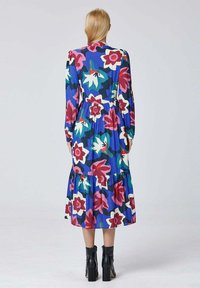 Une femme aux cheveux blonds droits porte une robe longue à manches avec un motif floral large en rose, blanc et vert, arrivant jusqu'aux mollets et des bottines noires.