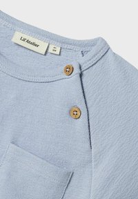 Lys blå kortermet topp med teksturert stoff, som har en liten brystlomme og to tresknapper på skulderen. Merkelappen er synlig.