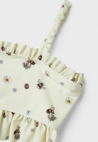 Top jaune à volants avec des motifs floraux et d'insectes, présentant des abeilles et des coccinelles, fabriqué en tissu extensible avec de fines bretelles réglables.