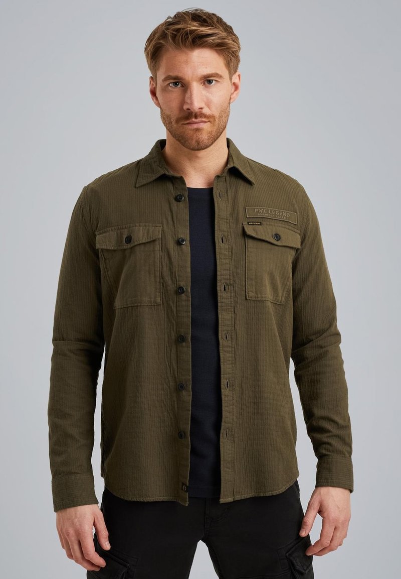 Camicia verde oliva a maniche lunghe in tessuto texturizzato. Presenta due tasche sul petto con pattine, un'etichetta nera sul lato destro e una vestibilità comoda.