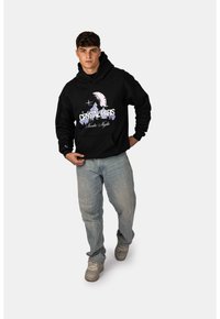 Černá mikina s kapucí s nápisem "CRYSTAL PARIS" a grafikou měsíce, přední kapsa, kombinovaná se světle modrými džíny a šedými teniskami.