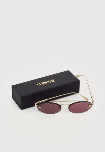 Οβάλ γυαλιά ηλίου με χρυσά πλαίσια και μοβ φακούς δίπλα σε μια μαύρη θήκη Versace με χρυσά γράμματα σε λευκό φόντο.