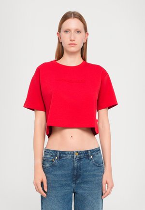 Punainen lyhythihainen cropped-t-paita, jossa on kohokuvioitu "MICHAEL KORS" -logo. Yhdistetty korkeavyötäröisiin sinisiin farkkuihin. Sujuva pintamateriaali.