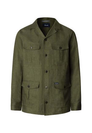 Junger Mann trägt eine olivgrüne, geknöpfte Jacke mit vier Klappentaschen über einem dunkelblauen Hemd und weißen Hosen, steht vor schlichtem Hintergrund.