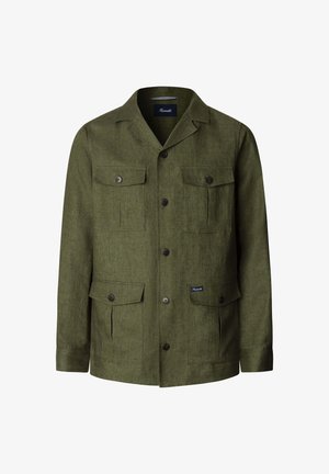 Joven con una chaqueta verde oliva abotonada con cuatro bolsillos con solapa, sobre una camisa azul marino y pantalones blancos, de pie frente a un fondo liso.