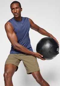 Haut de sport sans manches bleu marine avec une texture lisse, associé à un short vert olive, tenant un ballon médicinal noir avec une surface texturée.