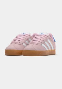 Par de sapatilhas Adidas Gazelle rosas com riscas brancas, solas de borracha, cordões rosas e tab's de calcanhar azuis, sobre fundo branco.
