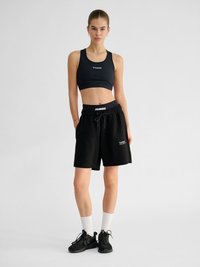 Jeune femme debout, portant un soutien-gorge de sport noir, un short noir avec cordon de serrage, des chaussettes blanches montantes et des baskets noires sur un fond clair uni.