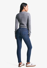 Siv volna pulover z rebrastimi manšetami, v kombinaciji s temno modrimi skinny kavbojkami. Obuti so v bele superge z rjavimi detajli, pogled od zadaj.