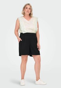 Kvinde iført ærmeløs hvid blonde top, sorte højtaljede shorts med lommer og hvide sneakers, stående med den ene hånd i lommen.