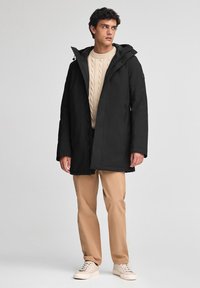 Parka nera impermeabile con cappuccio, zip anteriore e tasche laterali, indossata sopra un maglione crema a trecce e pantaloni beige.