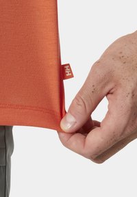 Oranžová košile vyrobená z hladké tkaniny, s visačkou na lemu. Ruka drží spodní okraj, čímž odhaluje čistý lem.