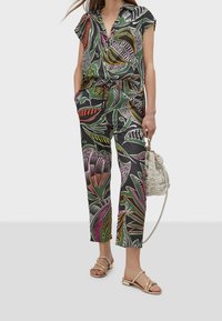 Květinový overal na krátký rukáv v zelené barvě, zdobený vícolorými vzory. Doplněný zlatými sandály a texturovanou béžovou taškou.