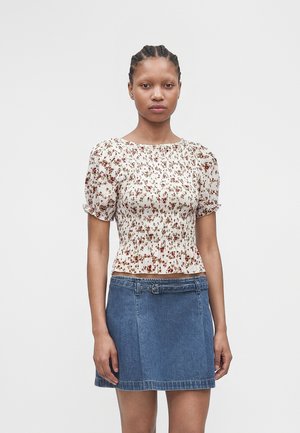 Frau mit geflochtenem Haar, die eine weiße Bluse mit Blumenmuster und Puffärmeln sowie einen blauen Jeansrock mit Gürtel trägt, steht vor einem schlichten Hintergrund.