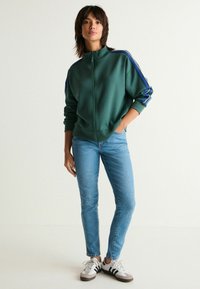 Vihreä vetoketjullinen treenitakki, jossa on siniset ja valkoiset sivuraidat, yhdistettynä vaaleansiniin slim-fit-farkkuihin ja valkoisiin tennareihin, joissa on mustia yksityiskohtia.
