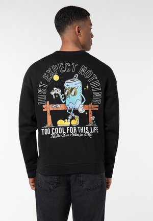 Schwarzer Sweatshirt mit einem farbenfrohen Grafik eines Cartoon-Getränkefigur mit Sonnenbrille, die auf einem Zaun sitzt. Der Text lautet: "ERWARTE EINFACH NICHTS."