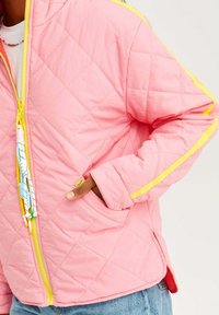 Prošívaná růžová bunda se žlutými akcenty, s uzavíráním na zip a ozdobnými třásněmi. Měkká textura a boční kapsy.