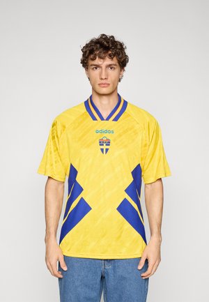 Żółta koszulka piłkarska z niebieskimi akcentami, z geometrycznym wzorem i niebieskim prążkowanym kołnierzem. Na przodzie logo Adidas oraz emblemat Szwecji.