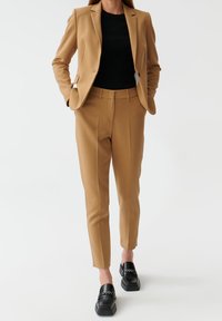 Blazer camel et pantalon assorti, associés à un haut noir côtelé et des chaussures noires à enfiler avec un détail chaîne. Tissu lisse, silhouette ajustée.