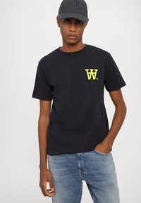 Schwarzes Baumwoll-T-Shirt mit kurzen Ärmeln und einem gelben Logo auf der linken Brust, kombiniert mit hellblauen Jeans.