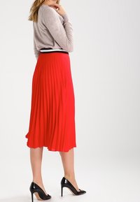Jupe midi plissée rouge avec une ceinture noire, associée à un haut manches longues gris clair et des escarpins noirs.