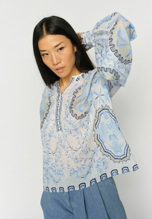 Woman with long black hair wearing a loose white blouse with blue floral patterns and blue pants, posing with one hand raised behind her head.