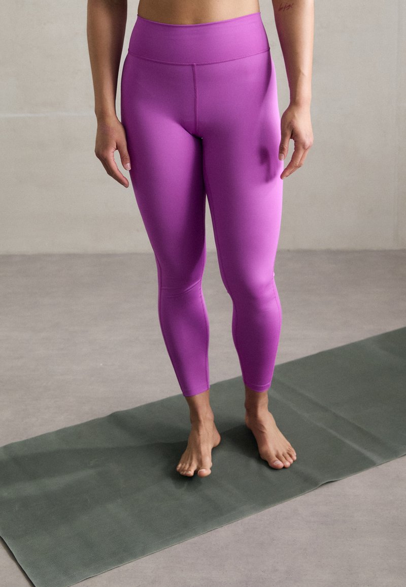 Leggings viola con vita alta, texture lucida e design aderente, abbinati a piedi nudi su un tappetino da yoga verde.