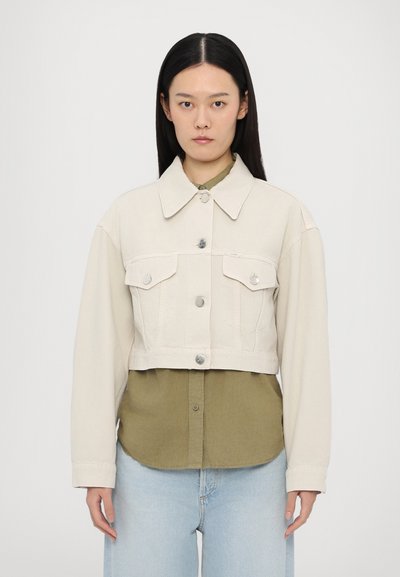 Beskåret beige denimjakke med to brystlommer og sølvknapper, lagdelt over en grøn skjorte, parret med lyseblå jeans.