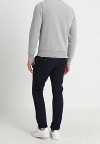Mann in grauem Langarm-Sweatshirt, dunklen Navy-Hosen und weißen Sneakers, der mit dem Rücken zur Kamera vor einem schlichten Hintergrund steht.