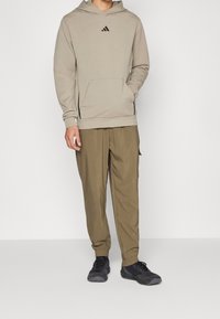 Felpa beige con cappuccio, tasca frontale e logo nero; abbinata a pantaloni cargo verde oliva e scarpe da ginnastica nere.