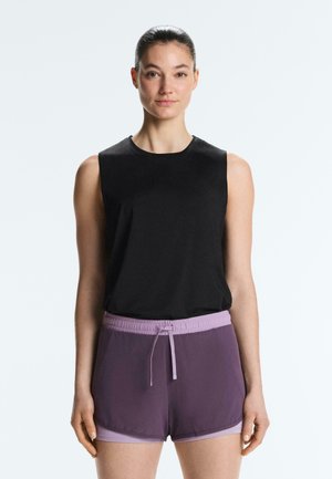 Femme aux cheveux foncés attachés en chignon porte un haut noir sans manches et un short de sport bicolore violet sur un fond clair uni.