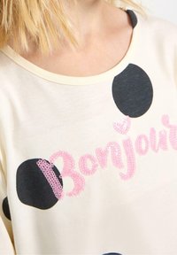 Hellblaus Baumwoll-T-Shirt mit schwarzen und marineblauen Punkten. "Bonjour"-Text in pinken Pailletten über der Brust, runder Ausschnitt, lässige Passform.