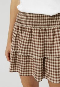 Jupe à volants à carreaux bruns et beiges avec taille élastique, portée avec un haut blanc, montrant une partie de la main et des jambes d'une personne.