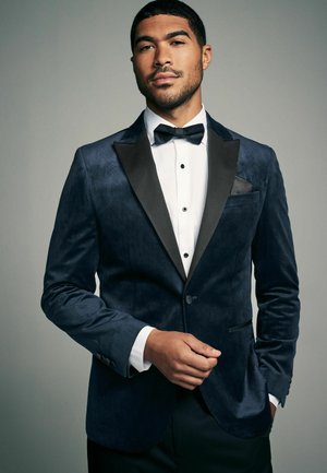 Giacca smoking in velluto blu navy con texture a motivi, rever in satin nero e chiusura a un bottone. Camicia bianca sotto con papillon nero.