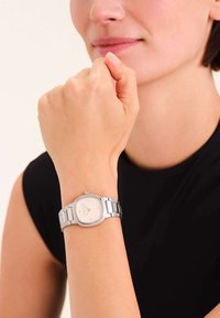 Reloj de acero inoxidable plateado con una esfera cuadrada blanca, números romanos y una pulsera de eslabones, llevado en una muñeca contra una blusa oscura.