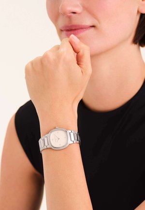 Orologio in acciaio inox argentato con quadrante bianco quadrato, numeri romani e bracciale a maglie, indossato su un polso contro una maglia scura.