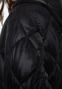 Sort pufferjakke med en skinnende, quiltet tekstur, der har diagonale syninger og en hætte. Materialet virker tykt og isoleret.