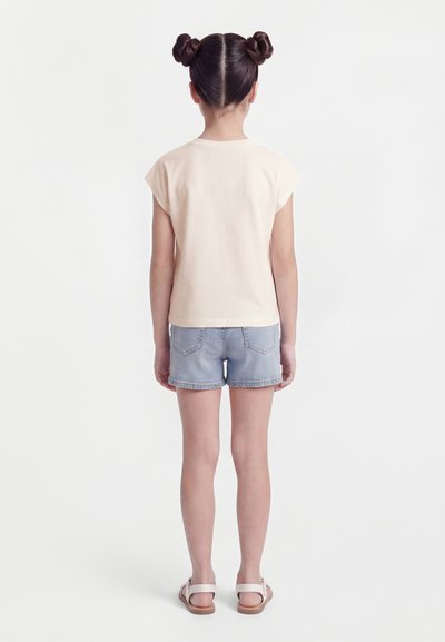 Enfant aux cheveux foncés coiffés en deux chignons, portant un t-shirt beige clair, un short en denim et des sandales blanches, debout de dos sur un fond uni.