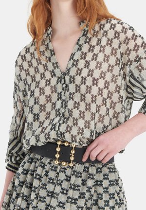 Femme portant un chemisier transparent à motifs et une jupe assortie avec une ceinture noire dotée d'une boucle en forme de boule dorée, main posée sur la taille.