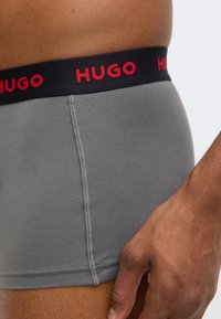 Grey cotton boxer shorts with a black elastic waistband featuring red "HUGO" lettering. The fabric appears smooth with visible stitching details.
