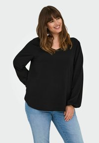 Crni dugi rukav s V-izrezom i opuštenim krojem, izrađen od mekog materijala, uparen s svijetloplavim trapericama. Jednostavan dizajn bez vidljivih uzoraka.