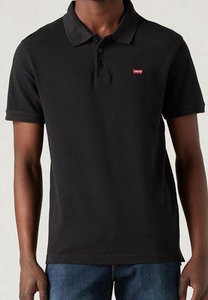 Homme portant un polo noir à manches courtes avec trois boutons et un petit logo rouge Levi's sur la poitrine, associé à un jean bleu.
