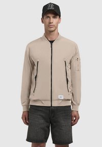 Beige bomberjakke med to forreste lynlås-lommer og en lomme på venstre arm, lavet af glat stof; parret med sorte shorts og en sort kasket.