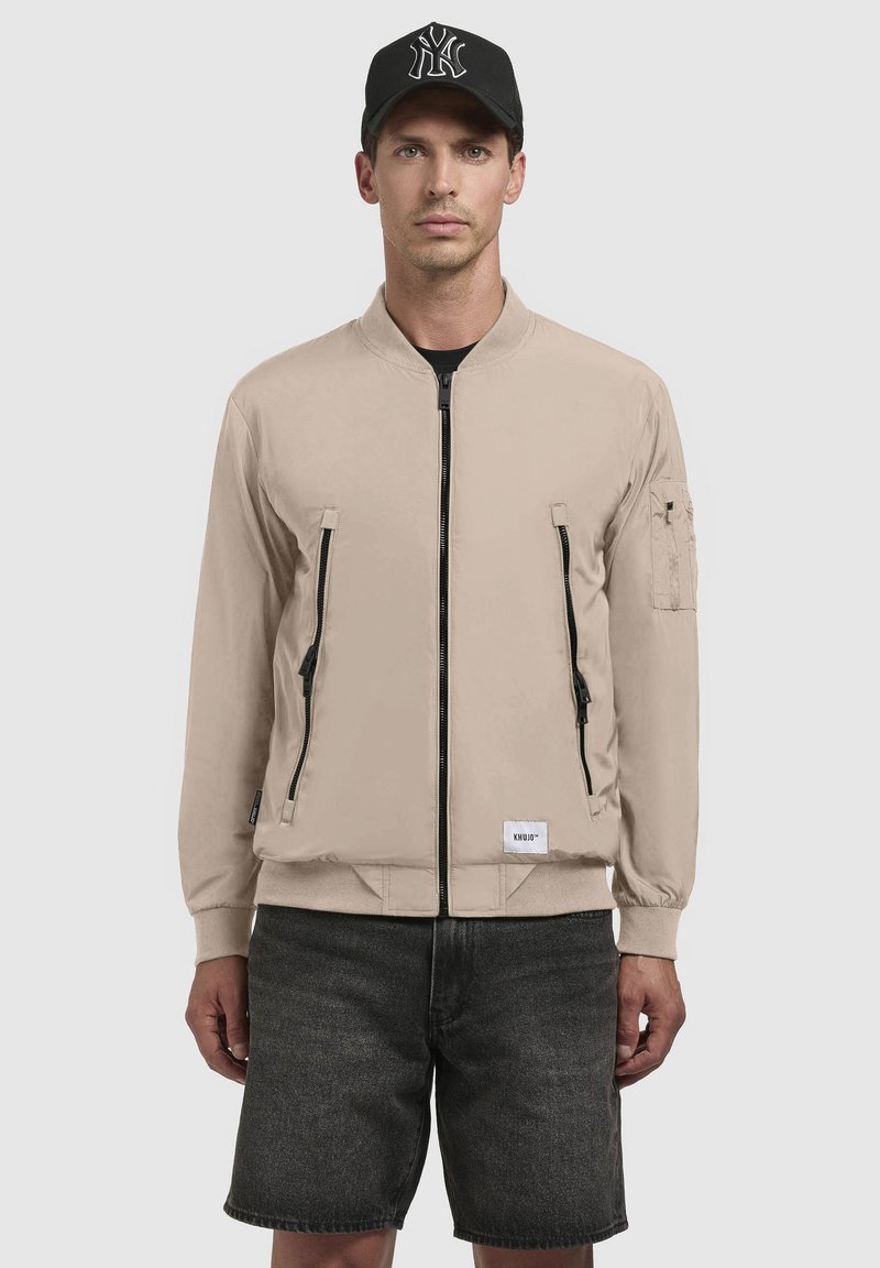 Beige bomberjack met twee voorzakken met rits en een zak op de linkerarm, gemaakt van gladde stof; gecombineerd met zwarte shorts en een zwarte pet.