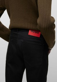 Brown ribbed sweater paired with black jeans. Visible red "HUGO" logo patch on waistband. Texture of sweater is knit, jeans are smooth.
