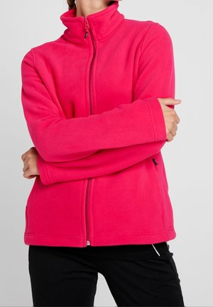 Person trägt eine leuchtend pinke Fleecejacke mit hohem Kragen und Frontreißverschluss, Arme vor der Brust verschränkt, vor einfachem Hintergrund.